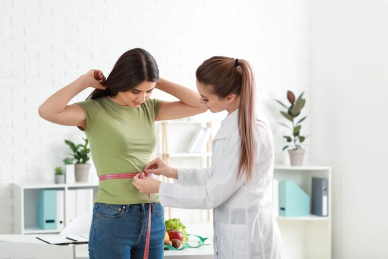 A doctor measuring a young woman’s waist with a tape measure during a weight loss evaluation in a medical office.