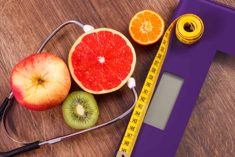 A purple weighing scale, stethoscope, measuring tape, and various fruits like grapefruit, apple, kiwi, and orange, representing a healthy approach to medical weight loss.