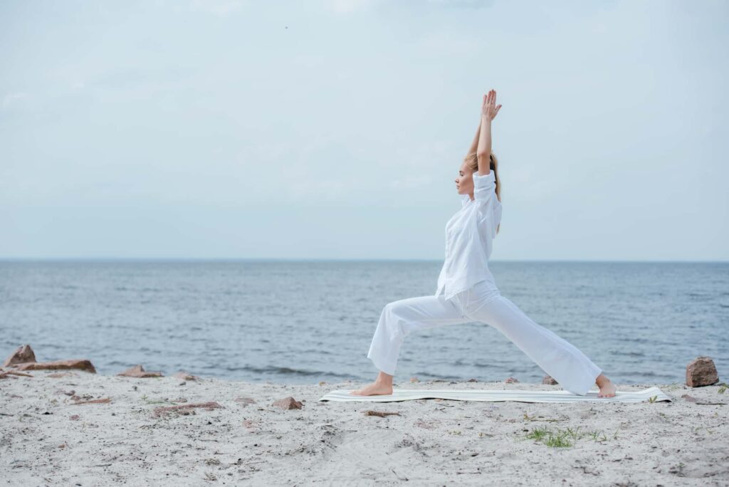 Woman practicing yoga in warrior pose on a serene beach, promoting mind-body balance and relaxation